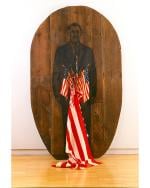 Wood plaque with a print of a man in a suit on it with six American flags plunged into his torso with one flag larger than the others, cascading down and pooling on the floor, similar to a stab wound and blood.