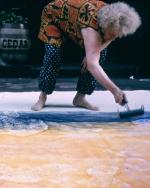 Elderly woman crouched over street with paint roller in hand, rolling blue color across webril laid over sidewalk in front of building with wooden barrel with the word "Cedar" printed on its side.
