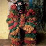 Figure in red, green, yellow raffia outfit and black mask. 
