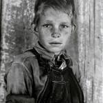 A blond boy in overalls and rolled up sleeves looking directly at the viewer.